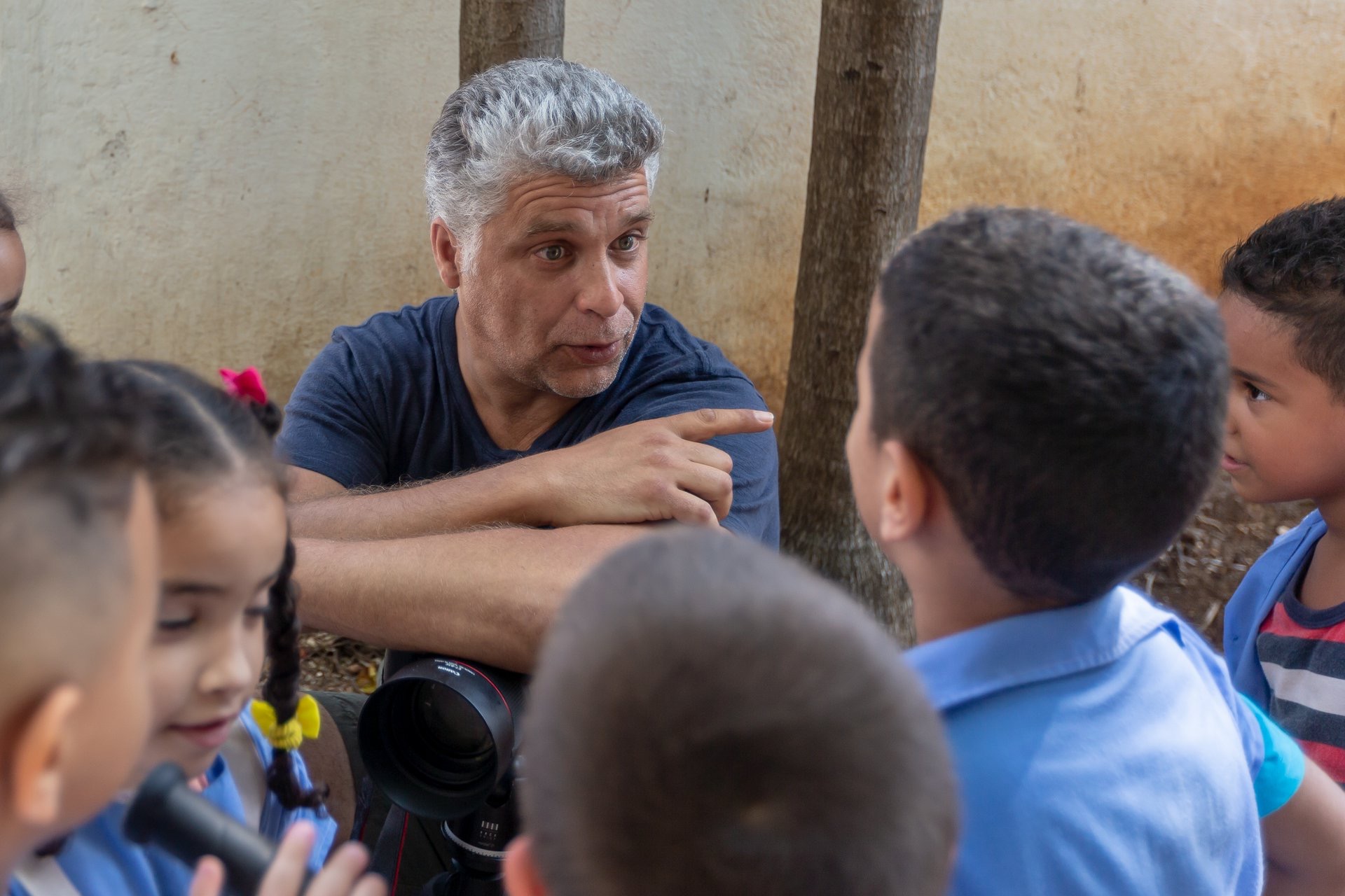 A candid outdoor shot of a gray-haired man in a blue shirt gesturing while speaking to a group of young children, conveying a mood of mentorship and engagement.
