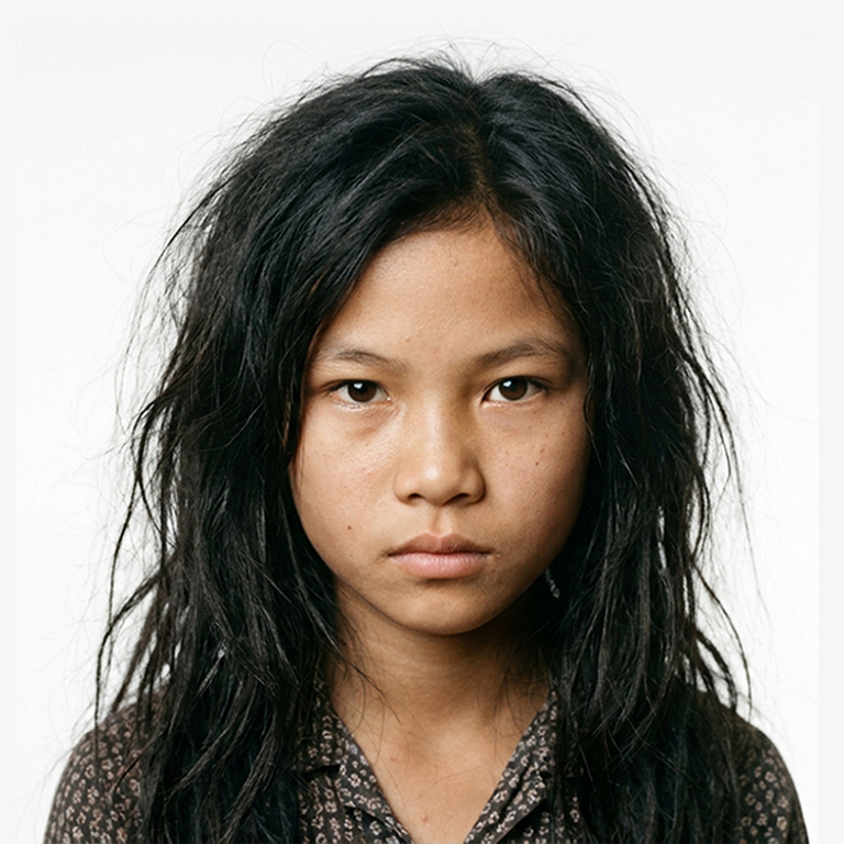 Sarah - a young orphan girl with long, wet dark hair and a serious, unflinching gaze, wearing a patterned collared shirt against a plain white background.